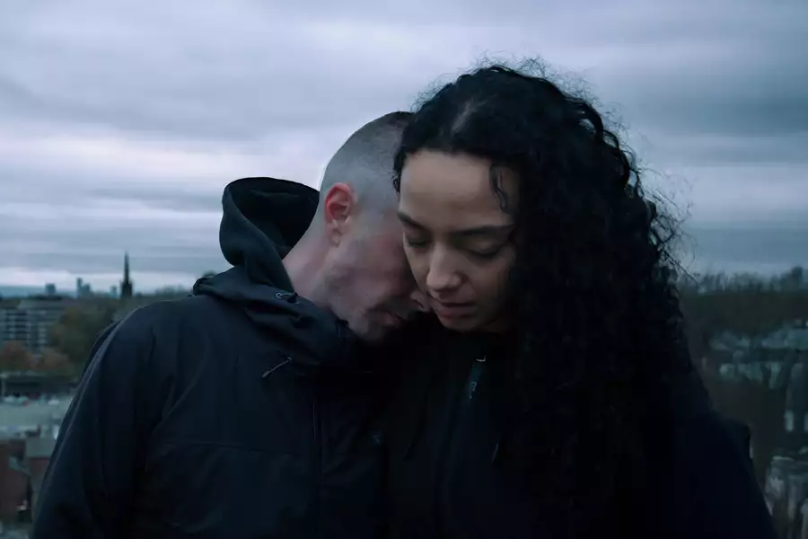 A man in a black hooded jacket leans his head against a woman’s shoulder, his eyes closed, while she looks downward with a solemn expression. The background shows an overcast sky and cityscape.