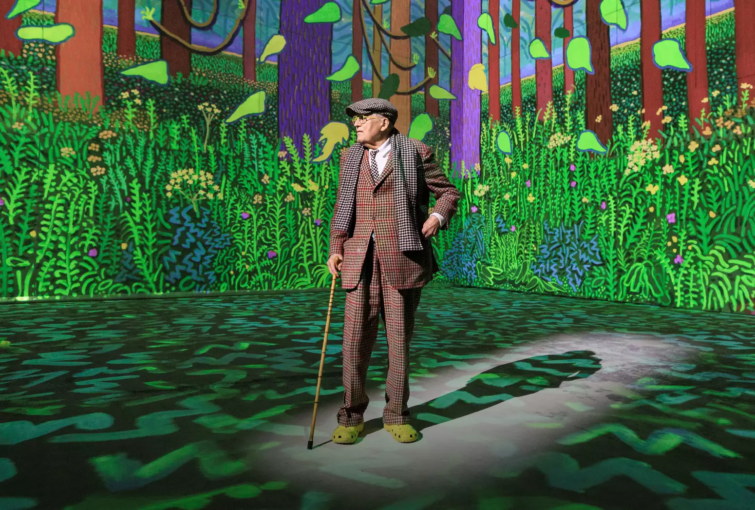 David Hockney wearing a chequered suit and yellow crocs – standing inside his exhibition Bigger & Closer (not smaller and further away) at Lightroom