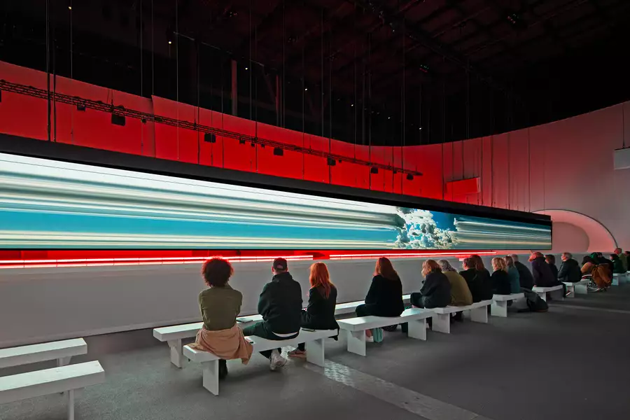 Spectators sit on a bench watching a long horizontal screen suspended over a white catwalk