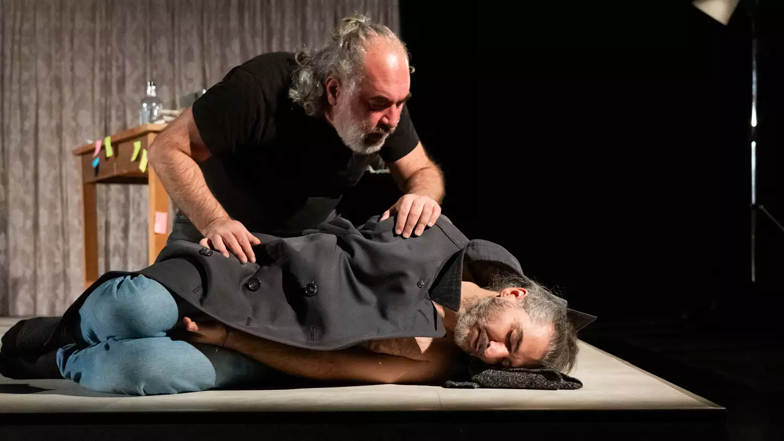A person lying down on stage with eyes closed underneath a coat – with another person peering over him