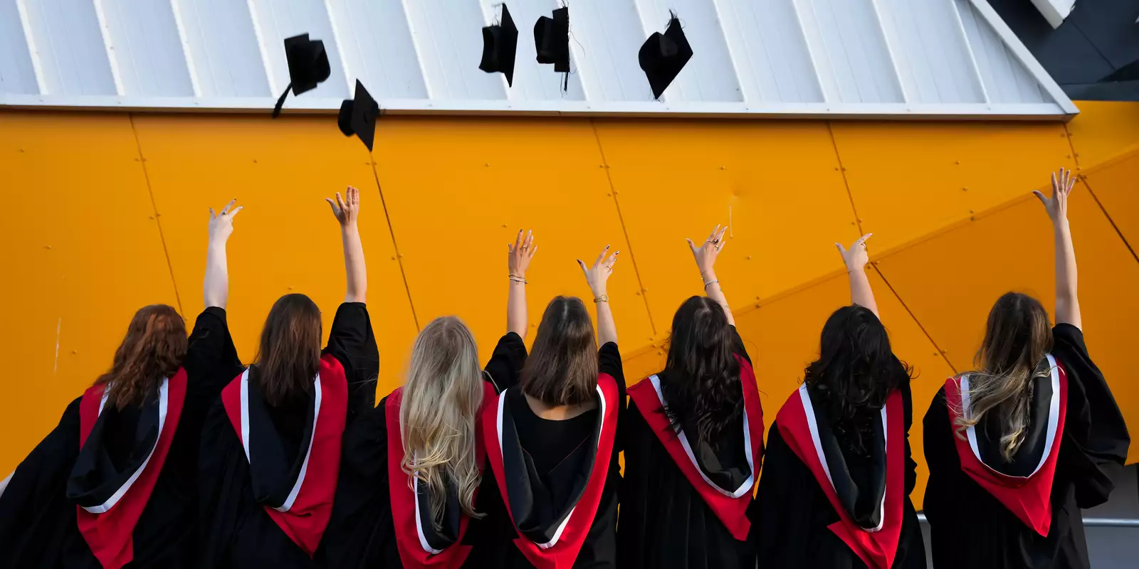 Graduates wearing gowns with their backs turned, throwing their hats in the air. They are stood outside Aviva Studios.