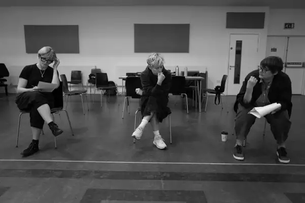 Black and white photograph of three people sat on a chair during THEY rehearsals