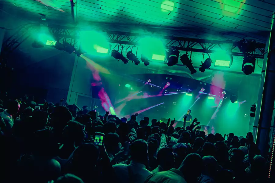 A DJ set on Festival Square from MIF23, showing a large crowd of people dancing and filming a DJ on stage. The lighting is hazy green and there are purple strobe lights.