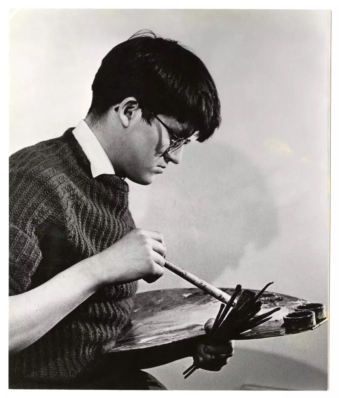 Black and white photograph of a young David Hockney holding an easel. He is wearing a jumper, shirt and glasses.