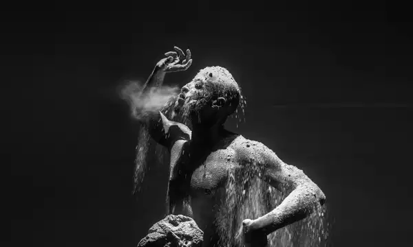 A dramatic black-and-white photograph of a man covered in dust and sand, with particles cascading over his body as he exhales forcefully.