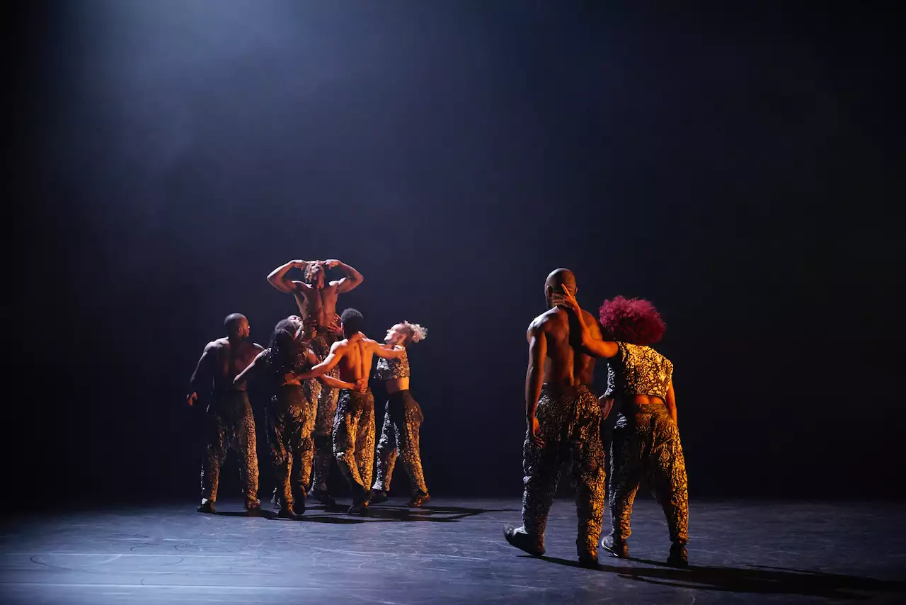 A group of contemporary dancers perform on a dimly lit stage, wearing patterned pants. One dancer is lifted above the rest with arms raised, while another pair interacts closely, creating a visually striking composition.