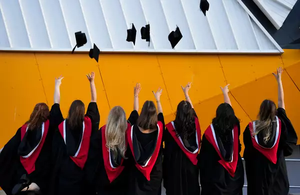 Graduates wearing gowns with their backs turned, throwing their hats in the air. They are stood outside Aviva Studios.