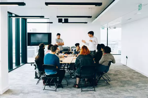 A number of people are sat around a large table while two people are stood speaking.