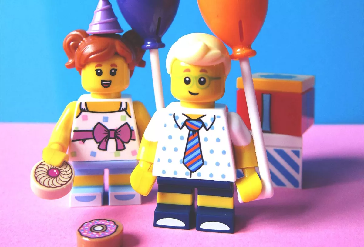 Two Lego mini-figures holding balloons and cake in front of wrapped presents