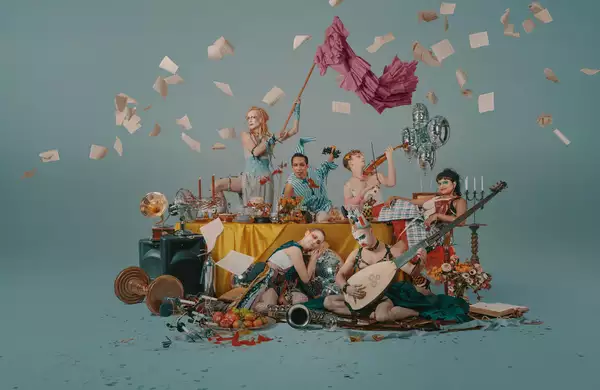 A group of performers around a table littered with objects, some are playing musical instruments, one is waving a flag