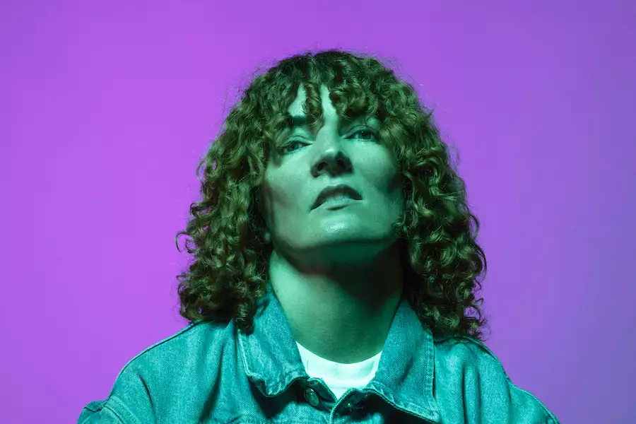 Electronic artist Planningtorock stands against a purple background wearing a denim jacket