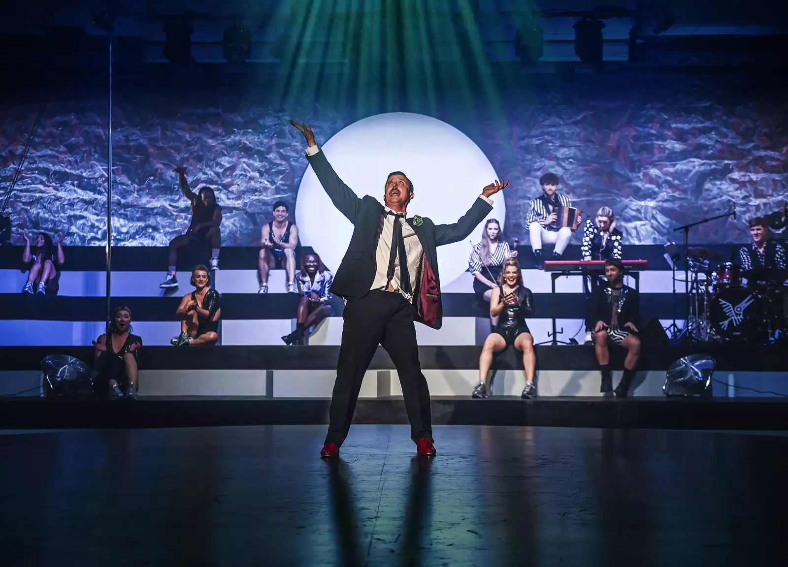 A performer dances in the middle of the stage wearing a suit during WAKE. Behind them, other performers sit on steps watching and clapping.
