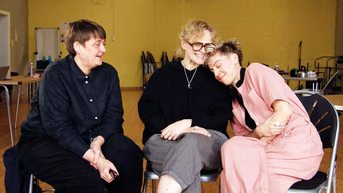 Maxine Peake, Sarah Frankcom and Imogen Knight sat on chairs smiling in a yellow rehearsal room