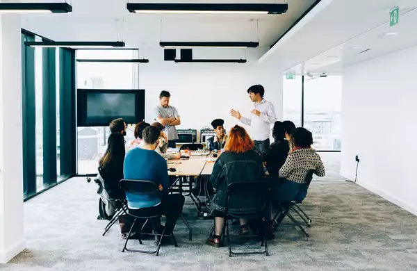 A number of people are sat around a large table while two people are stood speaking.