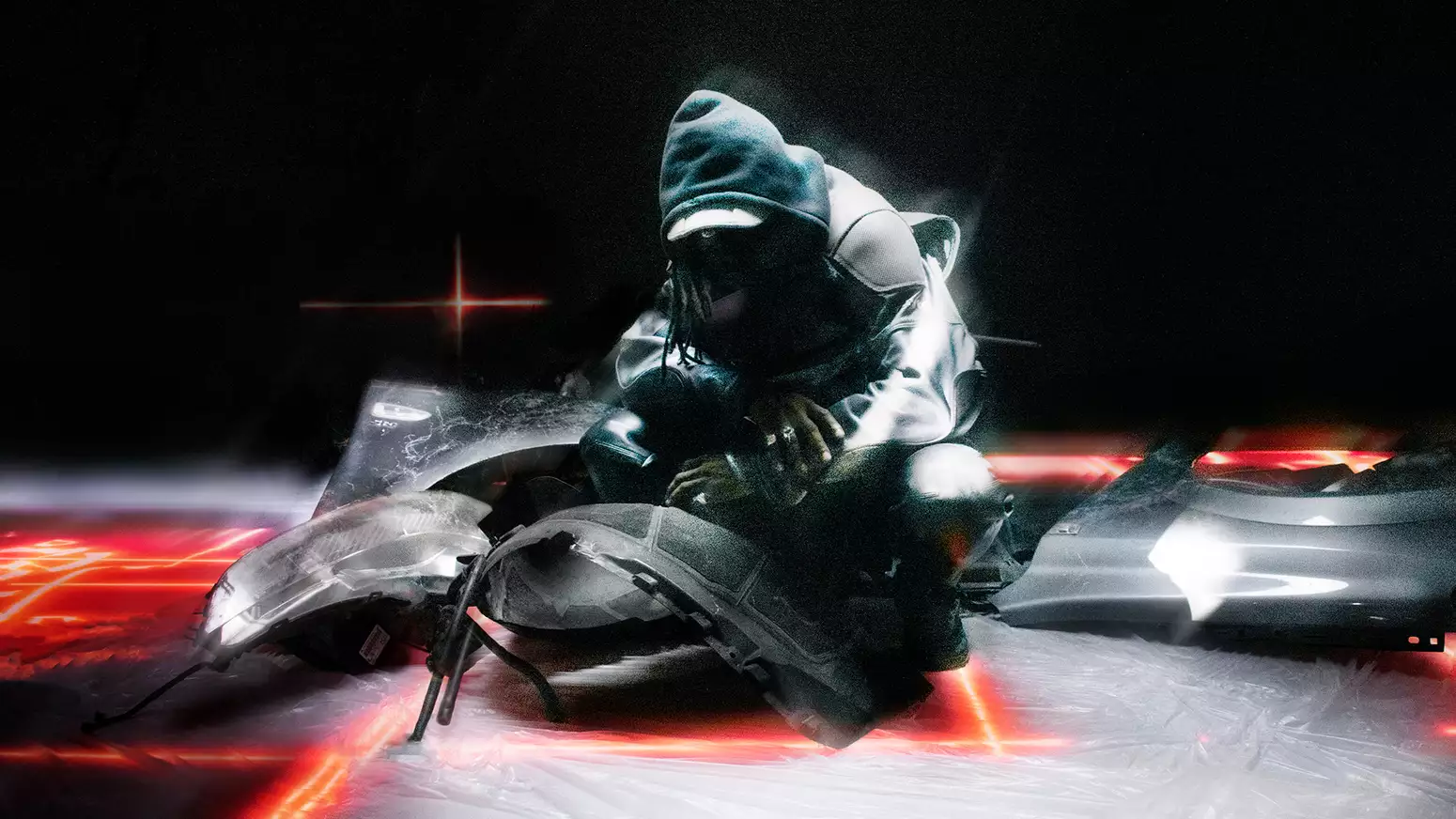 A person wearing all black with their hood up. They are crouching on the floor surrounded by fragments of a broken vehicle.