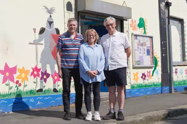 Three people stood on a curb outside smiling. Behind them is a wall decorated with paint and colourful drawings.
