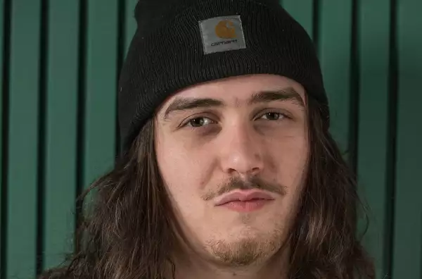A young man wearing a black Carhartt beanie stands in front of a green wall.