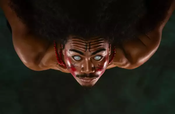 A member of FAFSWAG with glowing white eyes and red and black face paint stares intensely at the camera. They wear red beaded necklaces, and the image is flipped upside down.