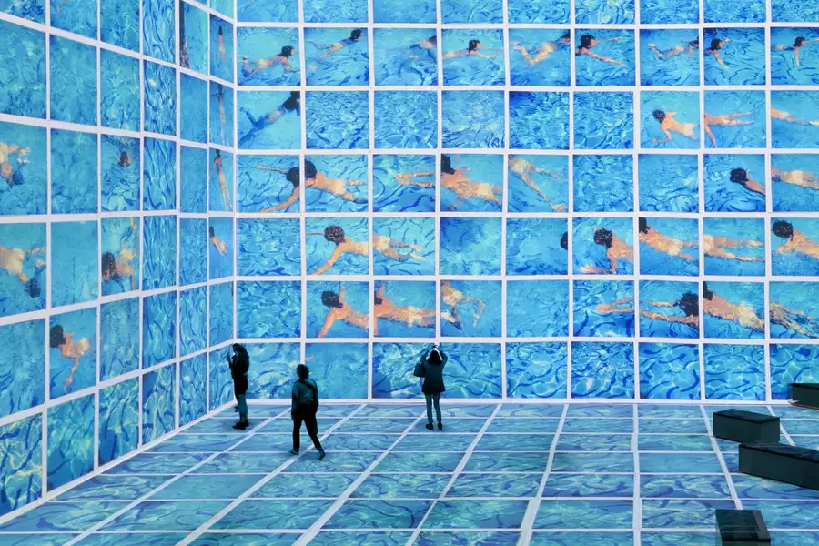 Three people stood in the David Hockney exhibition at Lightroom. His swimming pool paintings are shown on the screens.