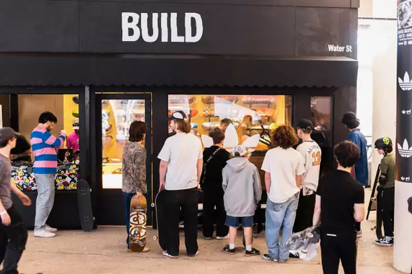 A queue of skateboarders waiting outside the Build Manchester pop-up skate shop, which is a black building with the word 'BUILD' on.