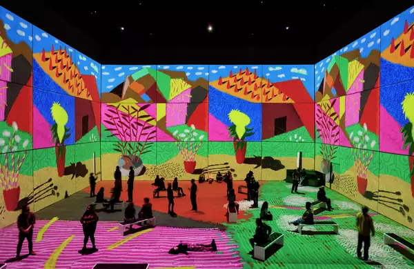 An image from David Hockney: Bigger & Closer (not smaller & further away), showing people in the exhibition seated and standing, surrounded by his artwork