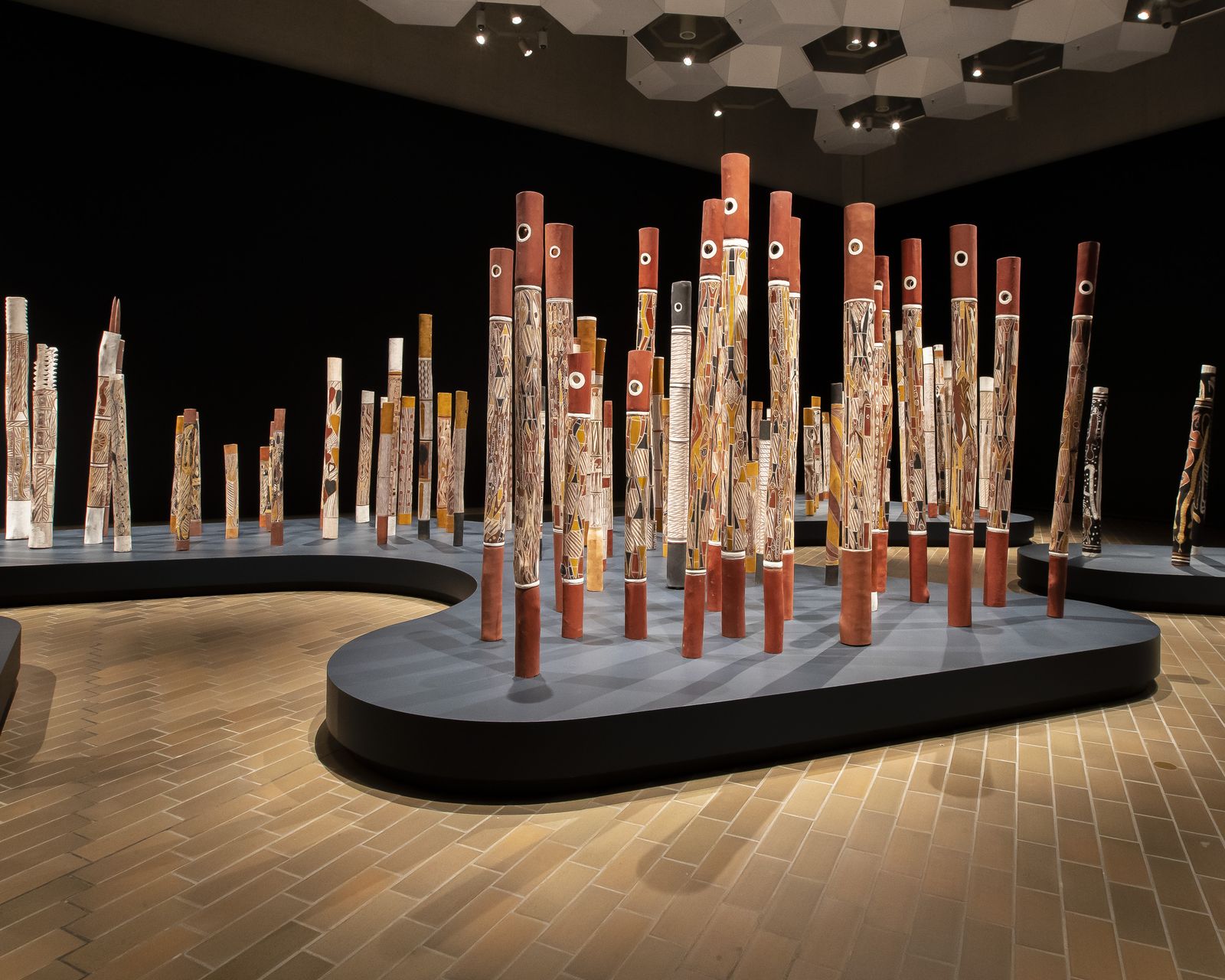 A photograph showing a section of an installation of 200 painted hollow logs standing upright in a large gallery space. The surrounding walls are painted in a dark colour and the logs are positioned on a navy plinth shaped to resemble both sides of a river bank.