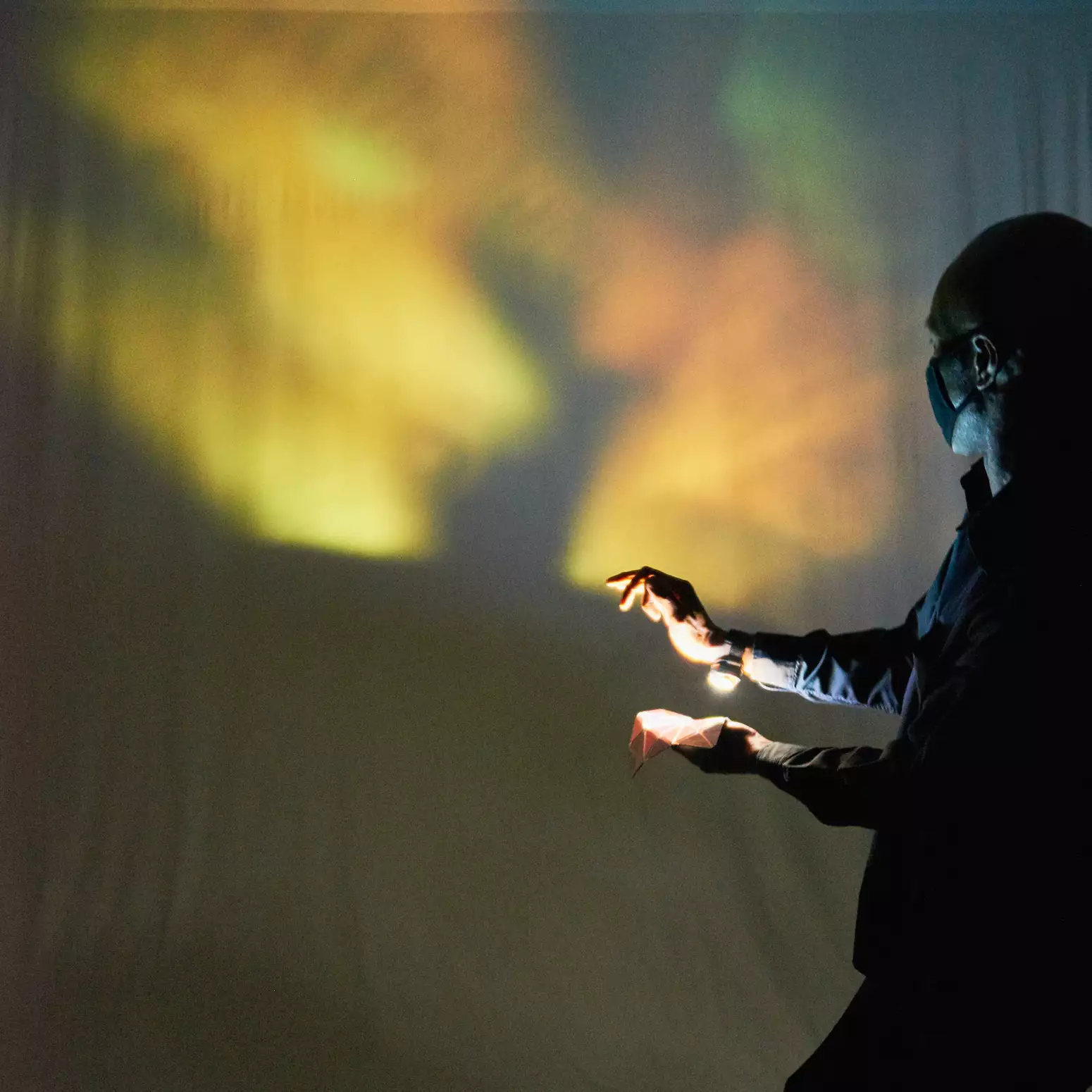 A performer on the far right of the image shines a light from their right wrist down at a kaleidoscope mirror matrix held up in their left hand. The yellow, orange and green reflected light from the kaleidoscope mirror matrix can be seen on the wall behind the performer; the shadow of their hands can also be seen.