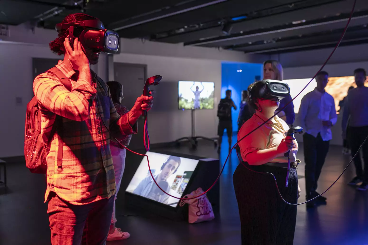 A man and a woman standing in a darkened art gallery, each wearing a Virtual Reality headset and holding a black controller. They are playing a game by the artist Rachel Maclean.