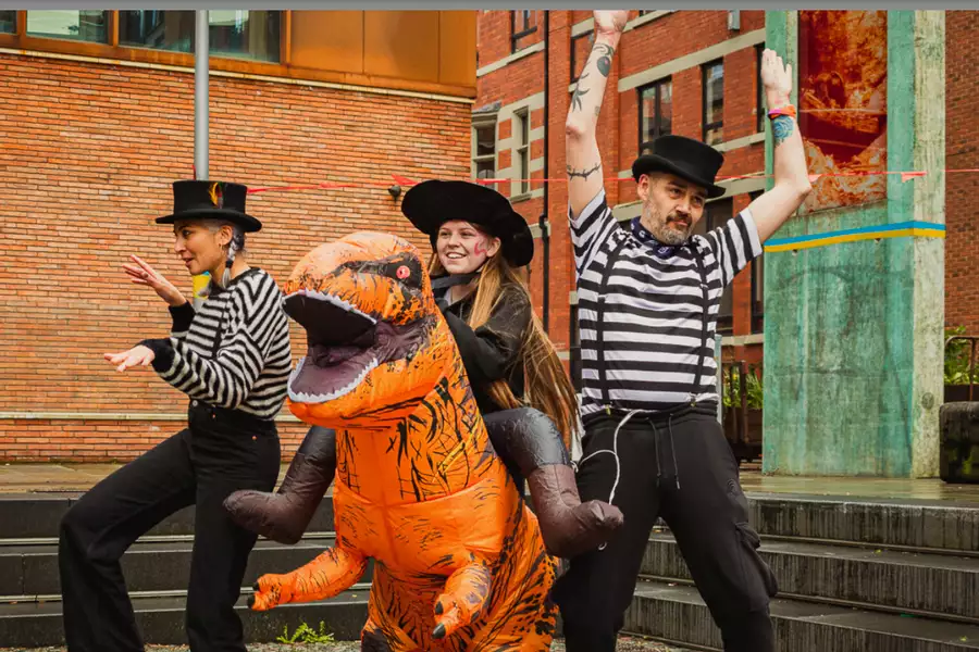 A person in an orange dinosaur costume, stood in between two people dressed in black-and-white striped tops.