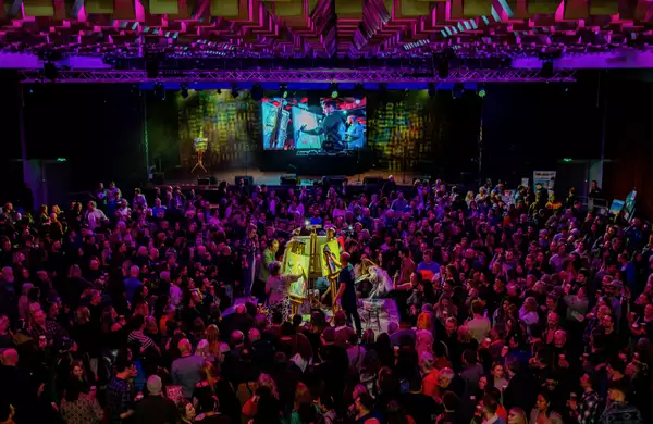 A large crowd gathered around a central stage featuring live painting, surrounded by colorful lighting and a dynamic ceiling design