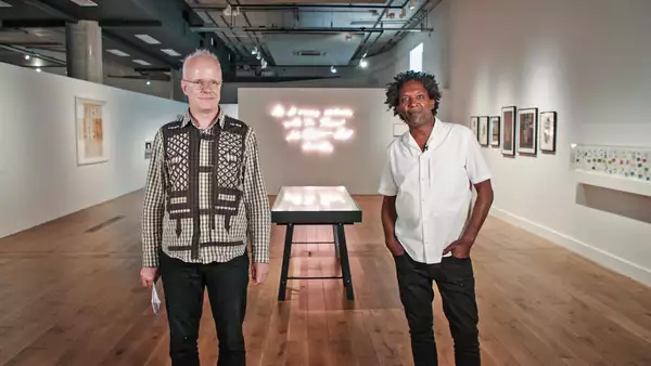 Hans Ulrich Obrist and Lemn Sissay stand in the Poet Slash Artist exhibition at HOME during MIF21. Hans Ulrich stands on the left; he is white with a pale peach skin tone and very short white hair; he wears glasses with clear frames, a green check shirt with black lattice and arrow patterns and dark navy jeans; he is smiling and looking just to the right of the camera. Lemn stands on the right; he is Black with a deep brown skin tone and medium length black and grey afro hair; he wears a white short sleeve shirt and black jeans, his hands are in the front pockets of his jeans; he smiles with a closed mouth directly at the camera. A vitrine can be seen between them in the background, and beyond that a neon artwork on the wall; a large frame hangs on the left wall behind Hans Ulrich; many smaller frames hang on the right wall behind Lemn. The industrial style ceiling of the space can be seen above the white gallery walls and partitions.