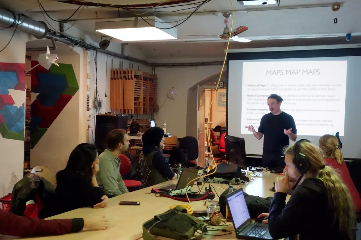 Josh Harle speaking to a group of people seated around a table in front of a slide presentation.