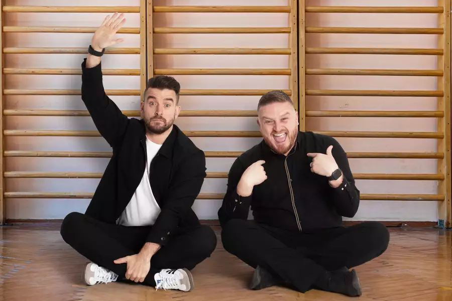 The Two Mr Ps sitting on the floor in front of wooden wall bars, one raising his hand and the other pointing at himself.