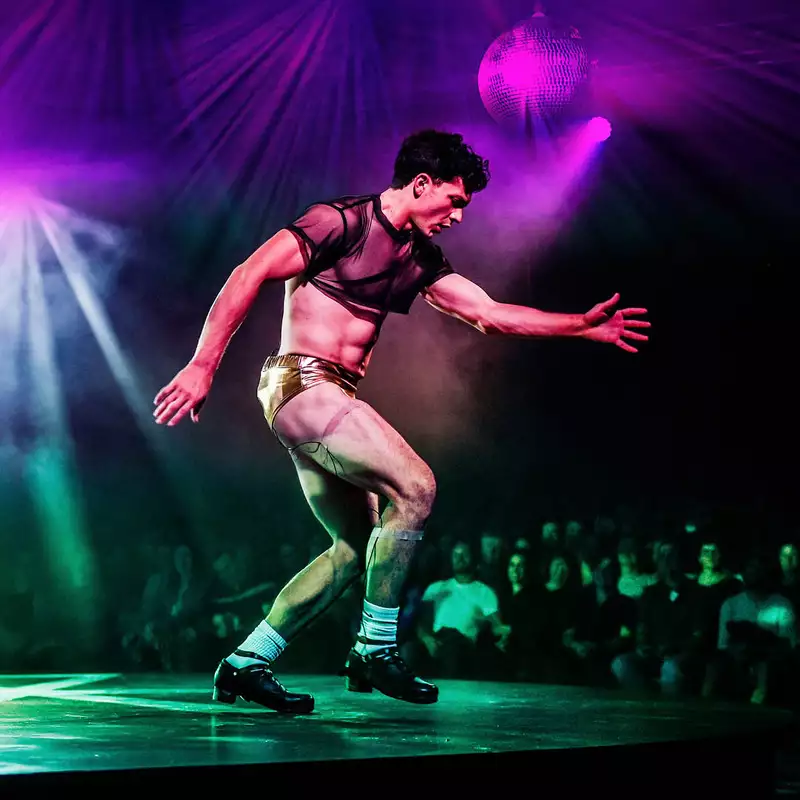 A dancer performs on stage during WAKE, wearing gold shorts and a sheer black crop top, illuminated by colorful stage lights.