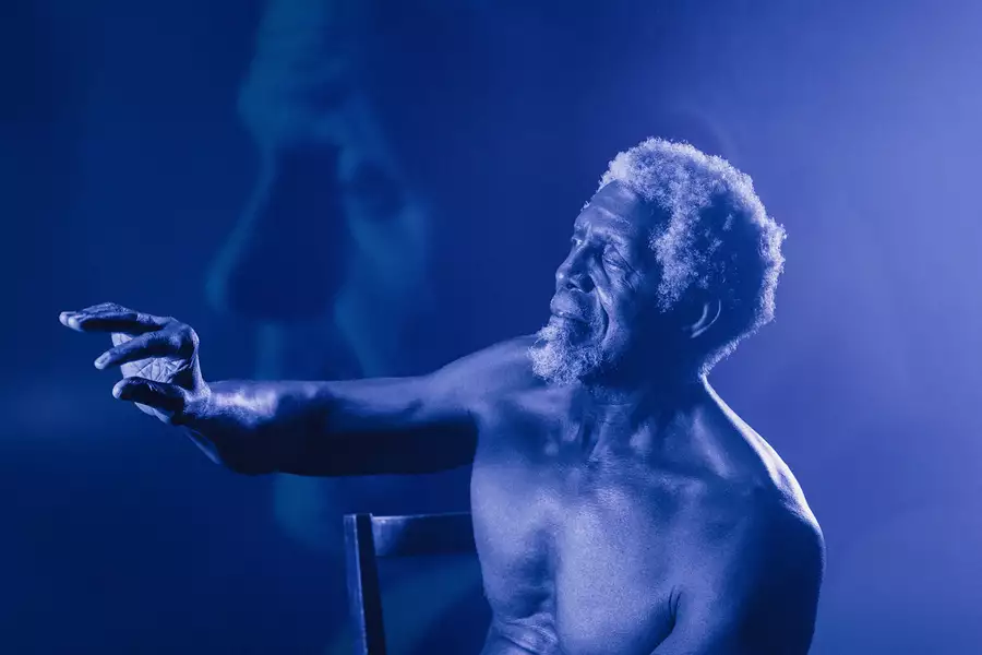 A shirtless elderly man with curly white hair and a beard sits on a chair, reaching out with an expressive hand. The image has a blue monochromatic tone with a soft-focus overlay of his face in the background.