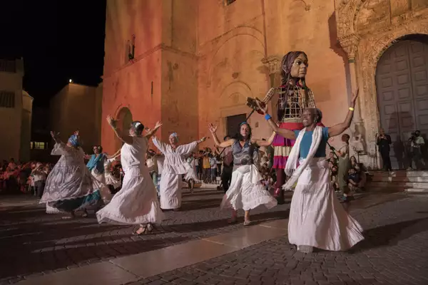 Performers in white skirts dance in front of a stone church lit up at night. Little Amal stands behind the dancers in front of the cathedral
