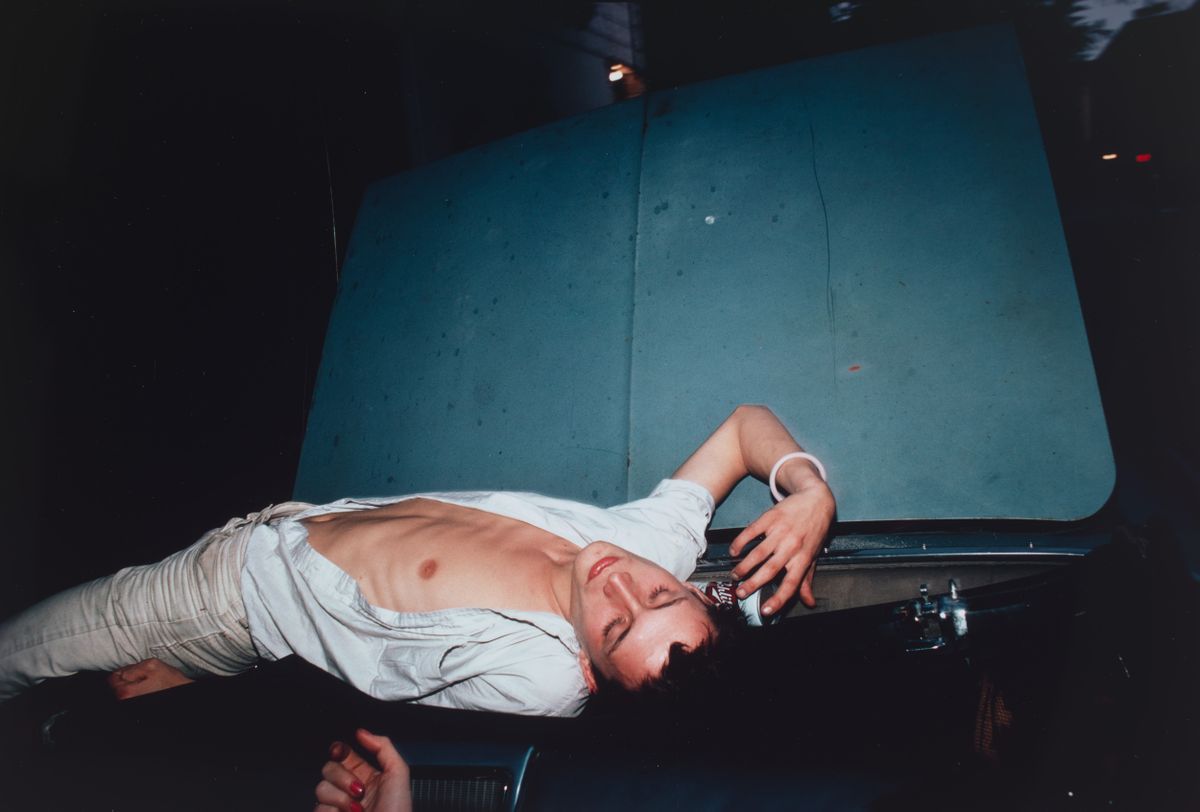 A male wearing a open shirt, laying on top of blue convertible car.