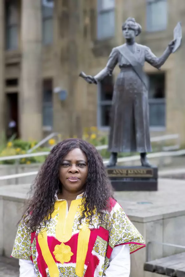 Headshot of Chantelle Tindall wearing a red and yellow patterned top. They are stood outside in front of a statue of the suffragette Annie Kenney.