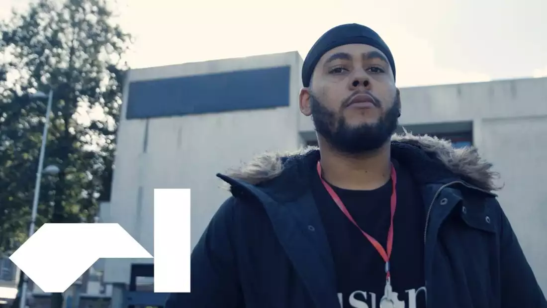 A young man with a beard looks towards the camera. He wears a black hooded jacket and red lanyard. The Factory International logo is visible in the bottom left corner.