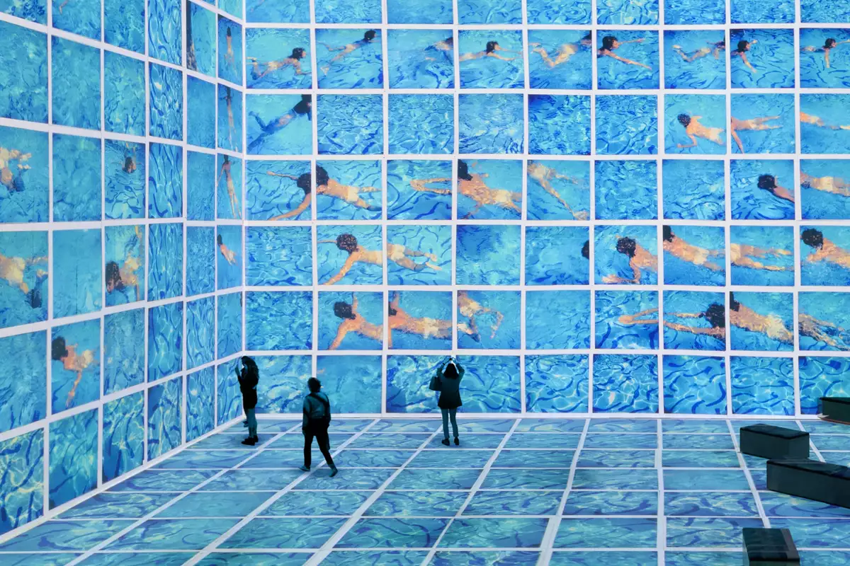 Three people stood in the David Hockney exhibition at Lightroom. His swimming pool paintings are shown on the screens.