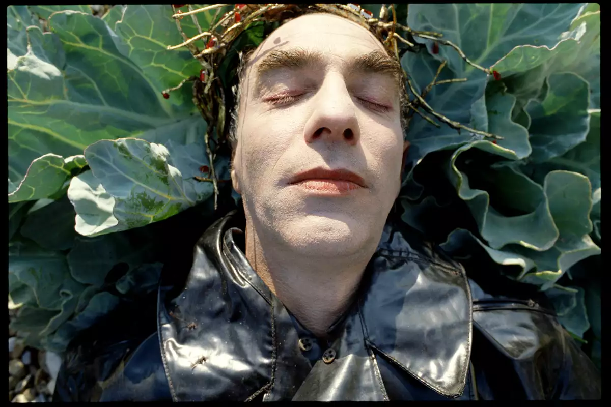 Still from Derek Jarman's The Garden (1990), showing a close up of Jarman's face. He is lying on a cabbage plant, wearing a black leather jacket and golden crown.