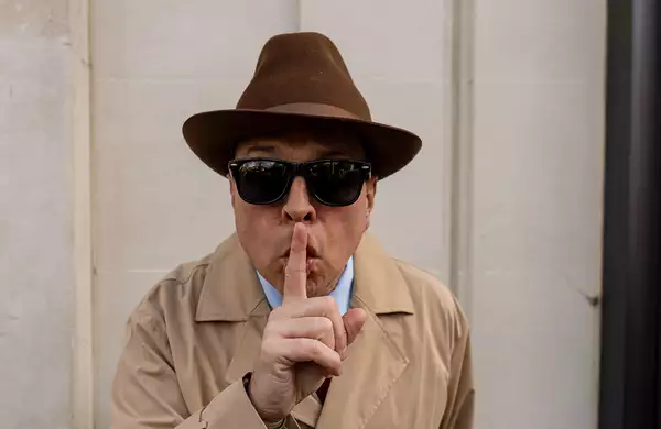 Dom Joly wearing a trench coat, fedora, and sunglasses gestures 'shush' with his finger to his lips."
