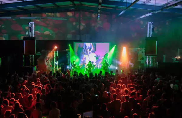 A live music performance in a dark indoor venue with colourful stage lighting and a large crowd enjoying the show. The stage features a projection screen displaying visuals, with green and orange lights illuminating the band.