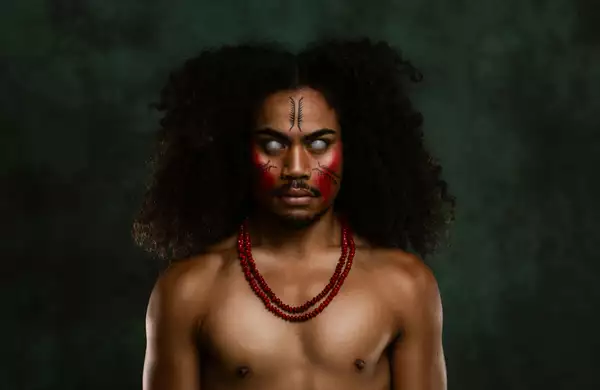 A member of FAFSWAG with glowing white eyes and red and black face paint stares intensely at the camera. They wear red beaded necklaces. The background is dark green.
