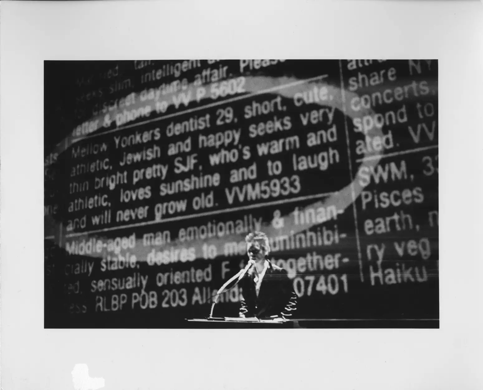 Black and white still from Laurie Anderson's United States Parts 1–4, showing Laurie talking into a microphone. Behind her is a large screen displaying a newspaper advert.
