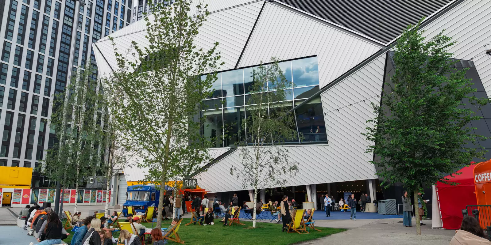 Groups of people sat outside Aviva Studios on the grass during MUBI FEST. There are yellow deck chairs plus a bar truck and coffee cart.