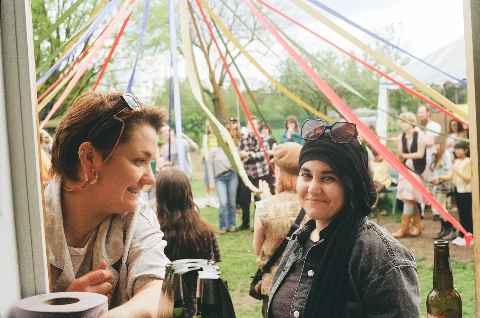 Two people smile and chat near a maypole decorated with colorful ribbons in an outdoor gathering, with other festival attendees in the background.