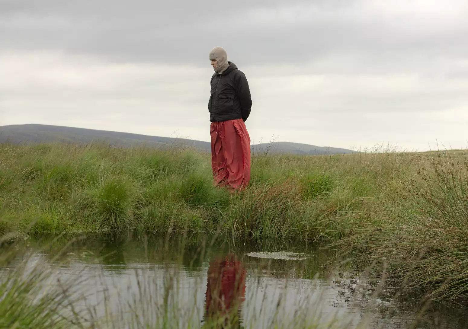 There is a pond in the foreground, a figure is stood in the surrounding reeds. It's a grey day. The lone person in the centre of the image wears a grey balaclava, black jacket and orange/red trousers. There are hills in the distance.