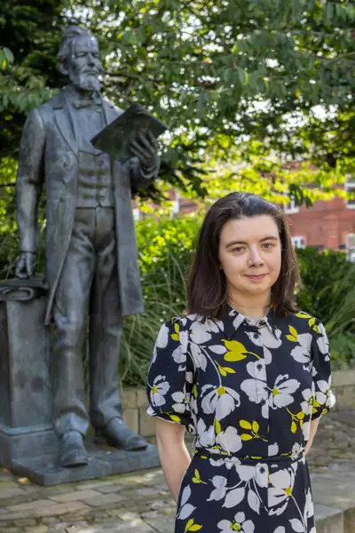 Headshot of Amelia Hall wearing a black dress with yellow and white flowers on it. They are stood outside in front of a statue and a tree.