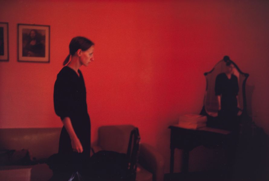 Cibachrome colour photograph of a woman standing in a red room, looking at her own reflection in a mirror